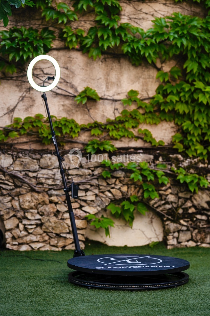 Plateforme circulaire avec un anneau lumineux sur pied, installée sur une pelouse devant un mur en pierre couvert de lierre.