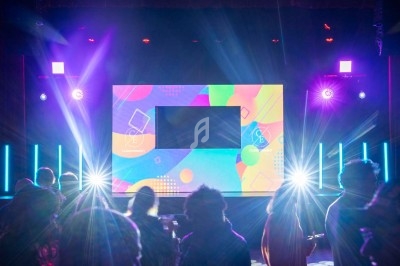Scène illuminée avec écran coloré au centre, éclairages vifs et silhouettes de spectateurs au premier plan.