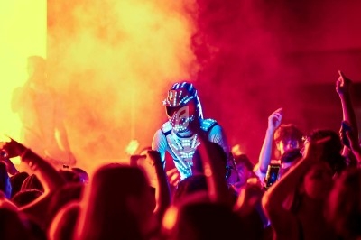 Un artiste en costume lumineux se produit au milieu d'une foule dans une ambiance colorée et enfumée.