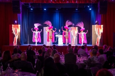 Des danseuses en costumes roses à plumes se produisent sur scène devant un public assis dans une salle éclairée.