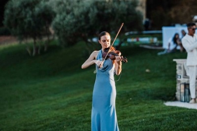 Une femme en robe bleue joue du violon sur une petite estrade en bois dans un jardin verdoyant.