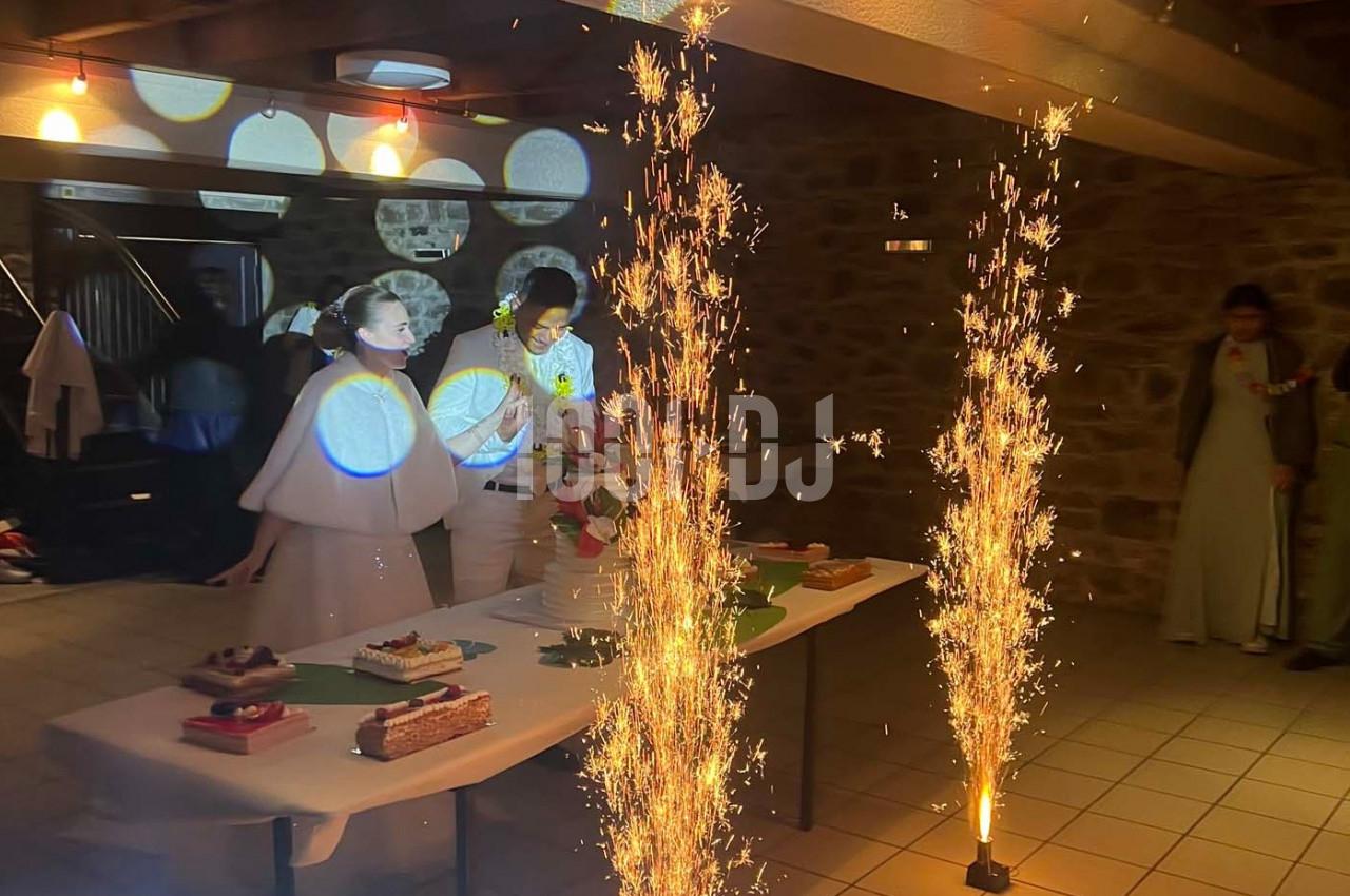 Un couple en tenue de mariage souffle des bougies sur un gâteau, entouré de décorations lumineuses et de fontaines…