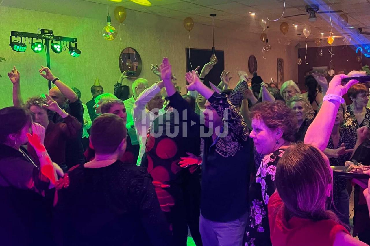 Des personnes dansent et lèvent les bras sur une piste éclairée par des lumières colorées lors d'une fête en intérieur.