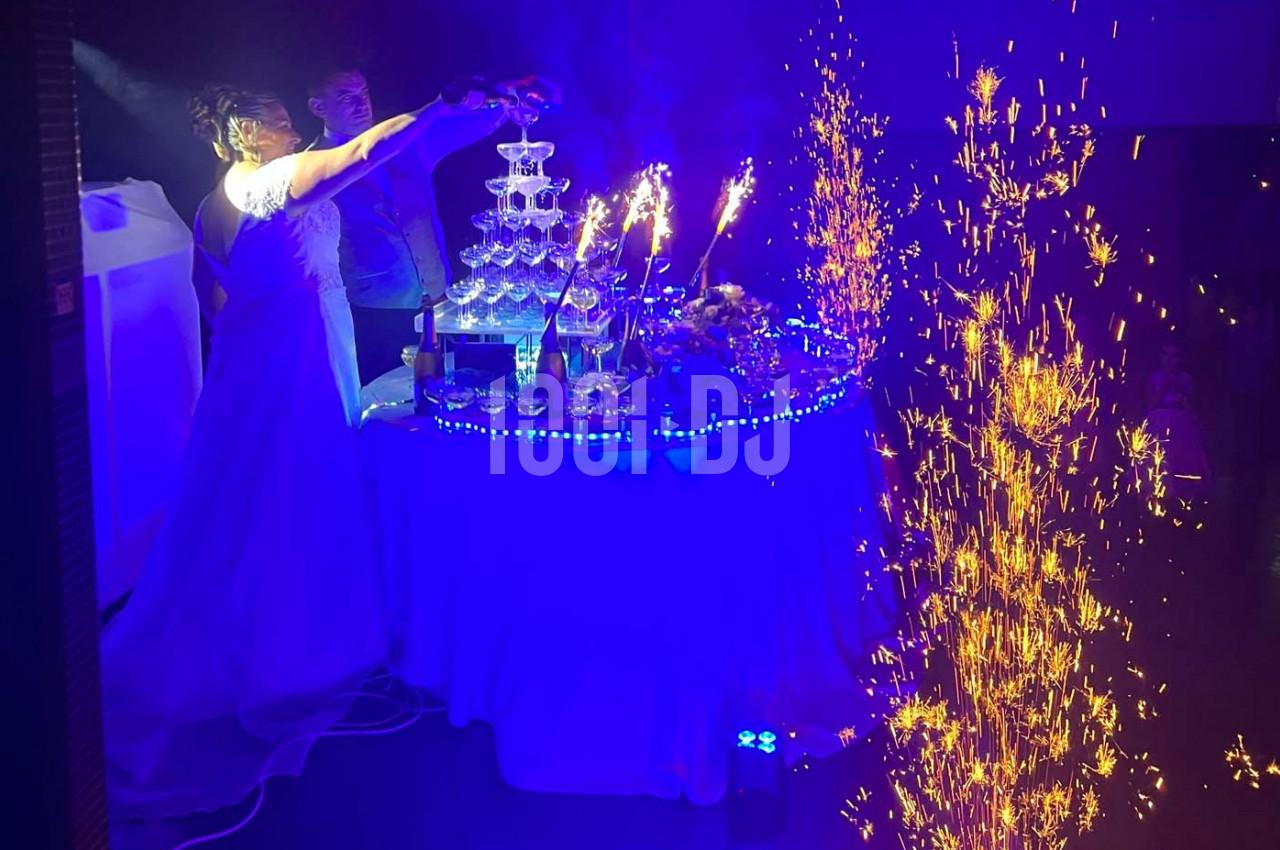 Une mariée verse du champagne dans une fontaine de verres illuminée, entourée de décorations et de gerbes d'étincelles.
