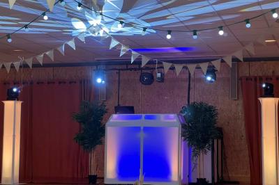 Scène décorée avec guirlandes lumineuses, plantes et une table de DJ éclairée en bleu et blanc.