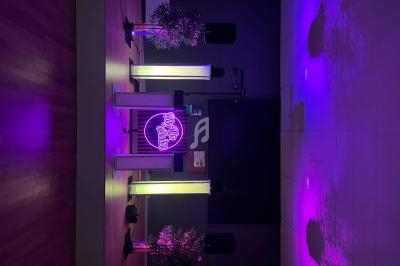 Salle sombre avec éclairage violet, colonnes lumineuses, plantes décoratives et enseigne néon au centre.