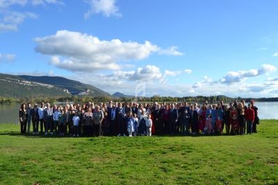 Un grand groupe de personnes posant sur une pelouse verte près d'un lac, avec des montagnes et un ciel bleu en arrière-plan.