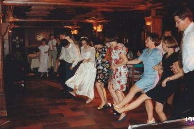 Des personnes, dont une mariée en robe blanche, dansent en ligne dans une salle au décor en bois.