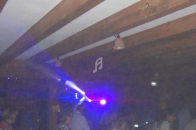 Salle sombre avec des poutres en bois, éclairée par des lumières colorées et des personnes debout ou en mouvement.