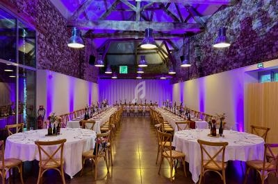 Salle de réception avec murs en pierre, poutres apparentes et tables rondes dressées sous un éclairage violet.