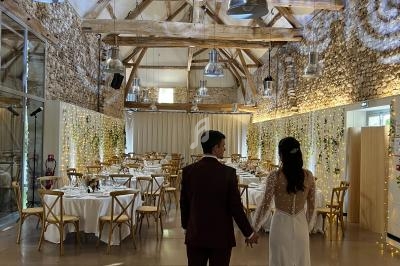 Un couple en tenue de mariage se tient la main dans une salle décorée avec des tables dressées et des lumières suspendues.