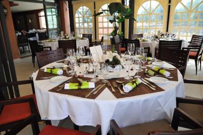Salle d'attente lumineuse avec fauteuils en rotin, coussins clairs, tables basses et plantes vertes sur un tapis rouge.