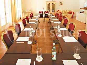 Salle d'attente lumineuse avec fauteuils en rotin, coussins clairs, tables basses et plantes vertes sur un tapis rouge.