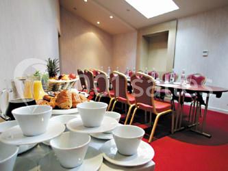 Salle d'attente lumineuse avec fauteuils en rotin, coussins clairs, tables basses et plantes vertes sur un tapis rouge.