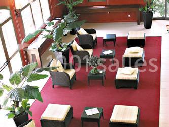 Salle d'attente lumineuse avec fauteuils en rotin, coussins clairs, tables basses et plantes vertes sur un tapis rouge.