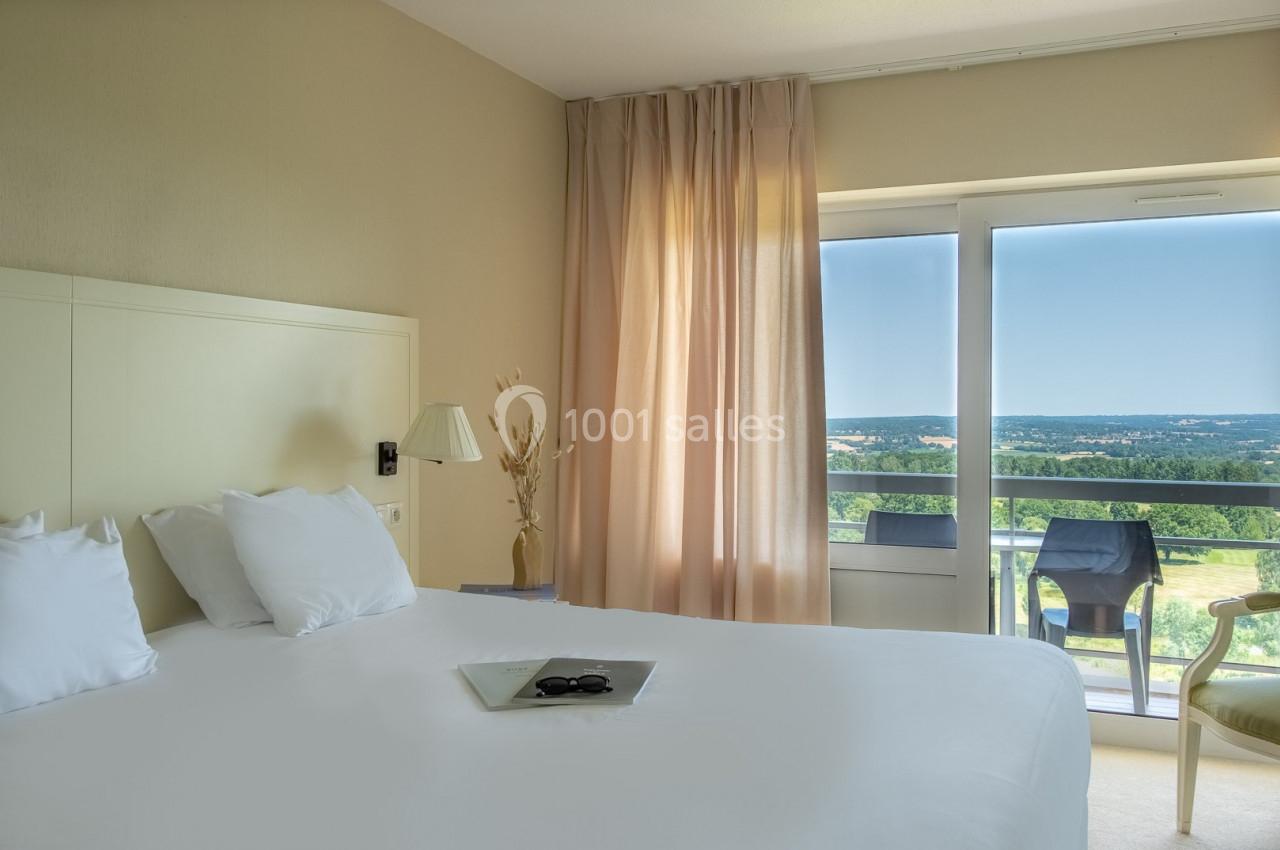 Chambre lumineuse avec un lit double, des rideaux beige clair et une vue sur un paysage verdoyant depuis un balcon.