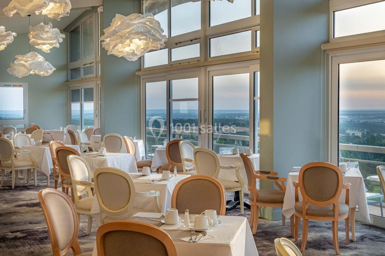 Salle de restaurant lumineuse avec grandes baies vitrées, tables dressées et vue sur un paysage extérieur.