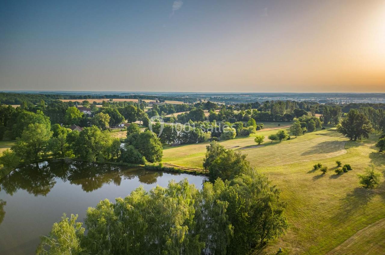 Paysage champêtre avec un étang entouré d'arbres, des prairies verdoyantes et un ciel dégagé au coucher du soleil.