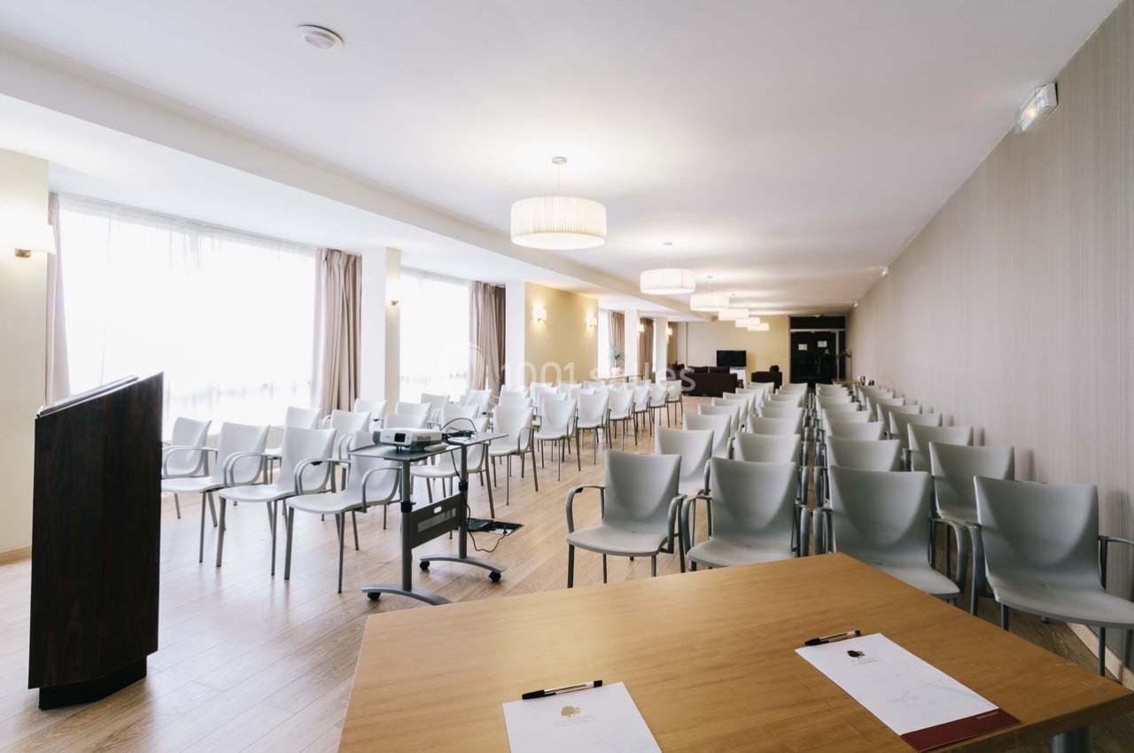 Salle de conférence lumineuse avec rangées de chaises, un pupitre et une table équipée de documents.