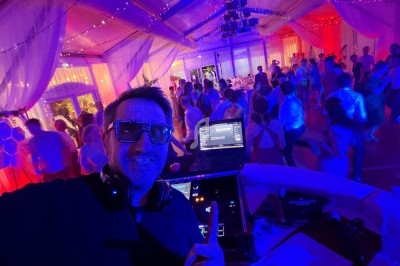 Un DJ souriant anime une soirée dansante sous une tente éclairée par des lumières colorées, avec des invités en arrière-plan.