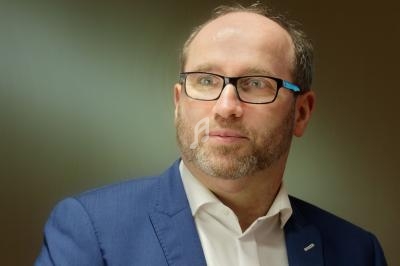 Homme portant des lunettes et un costume bleu, regardant légèrement sur le côté dans un environnement intérieur.