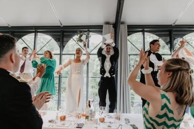 Des invités applaudissent un couple de mariés debout sur une table dans une salle lumineuse avec de grandes fenêtres.