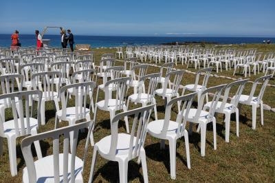 Chaises blanches disposées en rangées sur une pelouse près de la mer, avec quelques personnes en arrière-plan.
