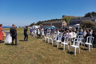 Un rassemblement en plein air avec des invités assis sur des chaises blanches, près de bâtiments et d'un paysage côtier.
