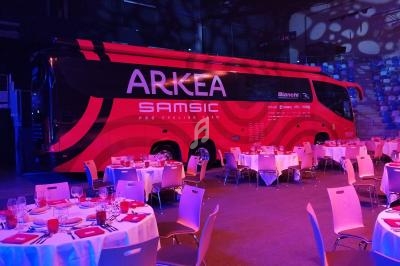 Autocar rouge de l'équipe cycliste Arkéa Samsic stationné dans une salle avec des tables dressées pour un événement.