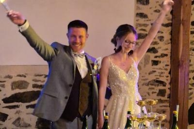 Un couple souriant en tenue de mariage tient des cierges magiques allumés près d'une pyramide de coupes de champagne.