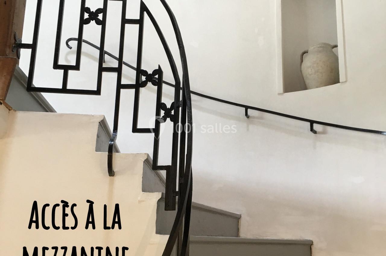 Escalier en colimaçon avec rampe en fer forgé menant à une mezzanine, mur blanc et niche décorative.
