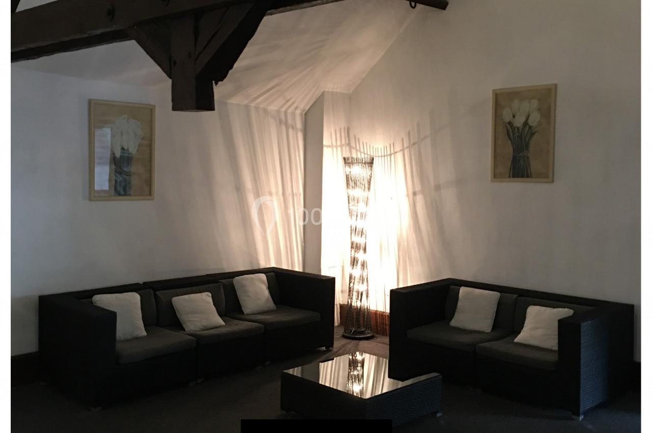 Deux canapés noirs avec coussins entourent une table basse dans un salon à poutres apparentes, éclairé par une lampe.