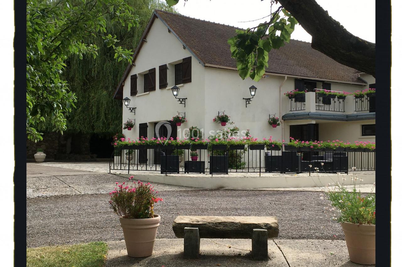 Maison traditionnelle avec volets en bois, entourée de fleurs, vue depuis un banc en pierre sous un arbre.