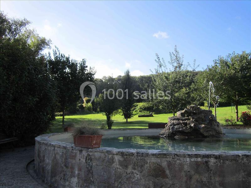Vue d'un jardin avec une fontaine en pierre au premier plan, entourée d'arbres et d'une pelouse sous un ciel dégagé.