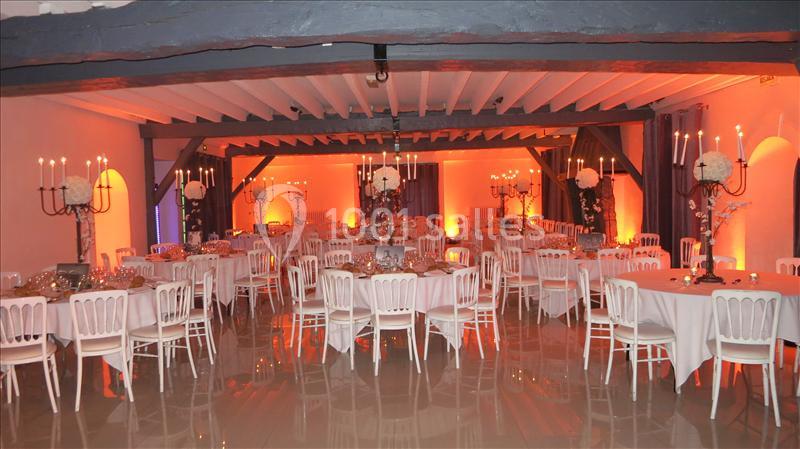 Salle de réception décorée avec des tables rondes, des chaises blanches et des chandeliers, éclairée par une lumière orangée.