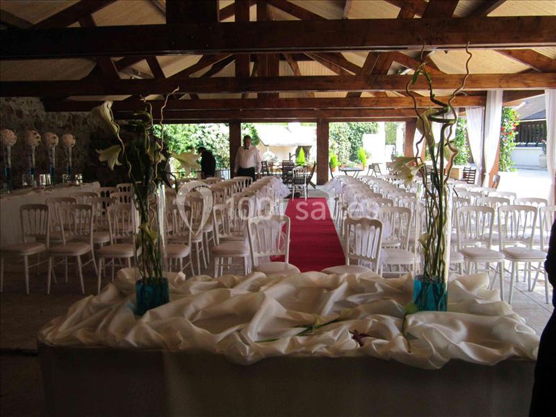 Salle décorée pour une cérémonie, avec chaises blanches alignées, tapis rouge central et compositions florales sur une table.