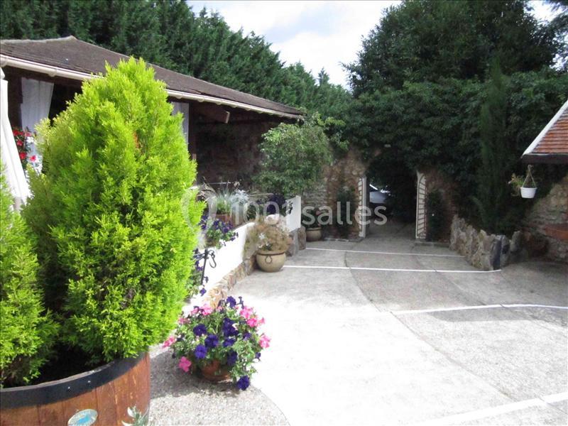 Cour extérieure avec plantes, fleurs colorées, murs en pierre et portail ouvert menant à un espace arboré.