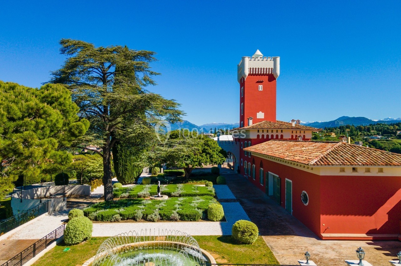 Vue d'un bâtiment rouge avec une tour, entouré de jardins, d'une fontaine et de végétation sous un ciel bleu.