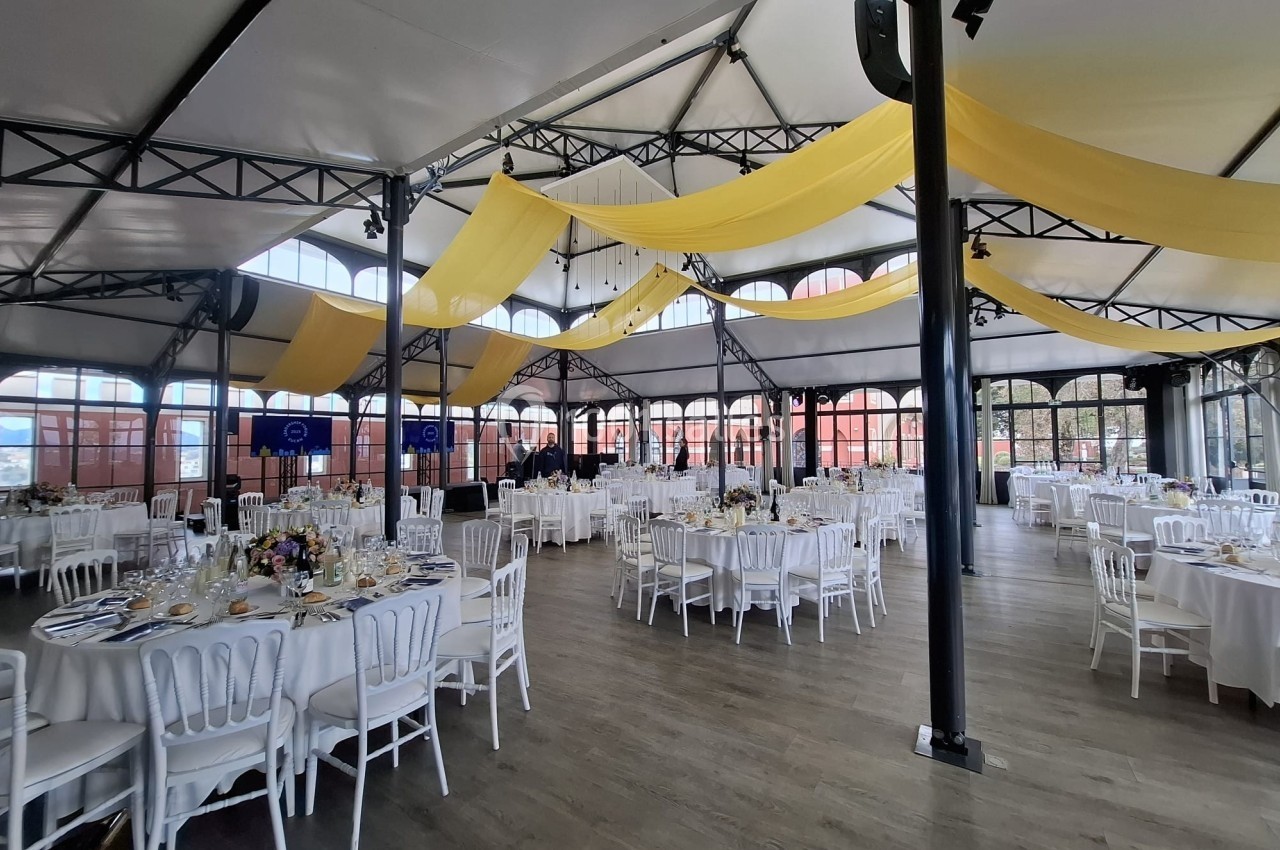 Salle de réception lumineuse avec tables rondes dressées, chaises blanches et décorations jaunes suspendues.
