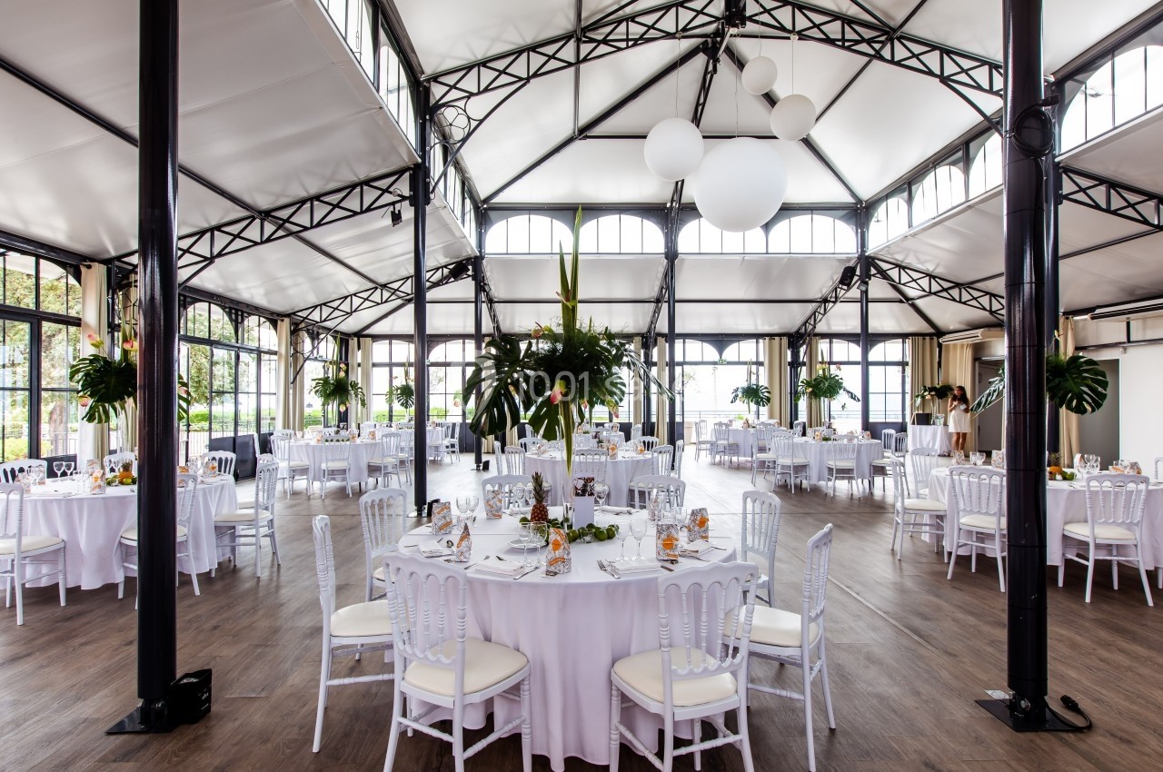 Salle de réception lumineuse avec tables rondes dressées, chaises blanches et décorations végétales.