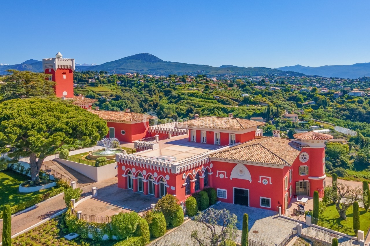 Vue aérienne d'une grande villa rouge entourée de jardins, située dans une région vallonnée et verdoyante.