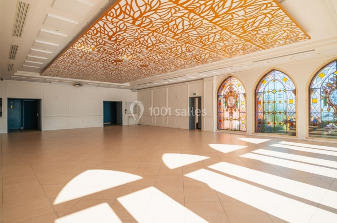 Salle spacieuse avec un plafond décoratif doré, des vitraux colorés et un sol carrelé éclairé par la lumière naturelle.