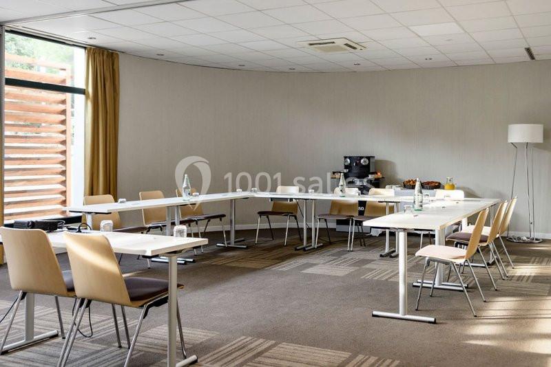 Salle de réunion lumineuse avec tables disposées en U, chaises en bois clair et coin avec machine à café et collations.