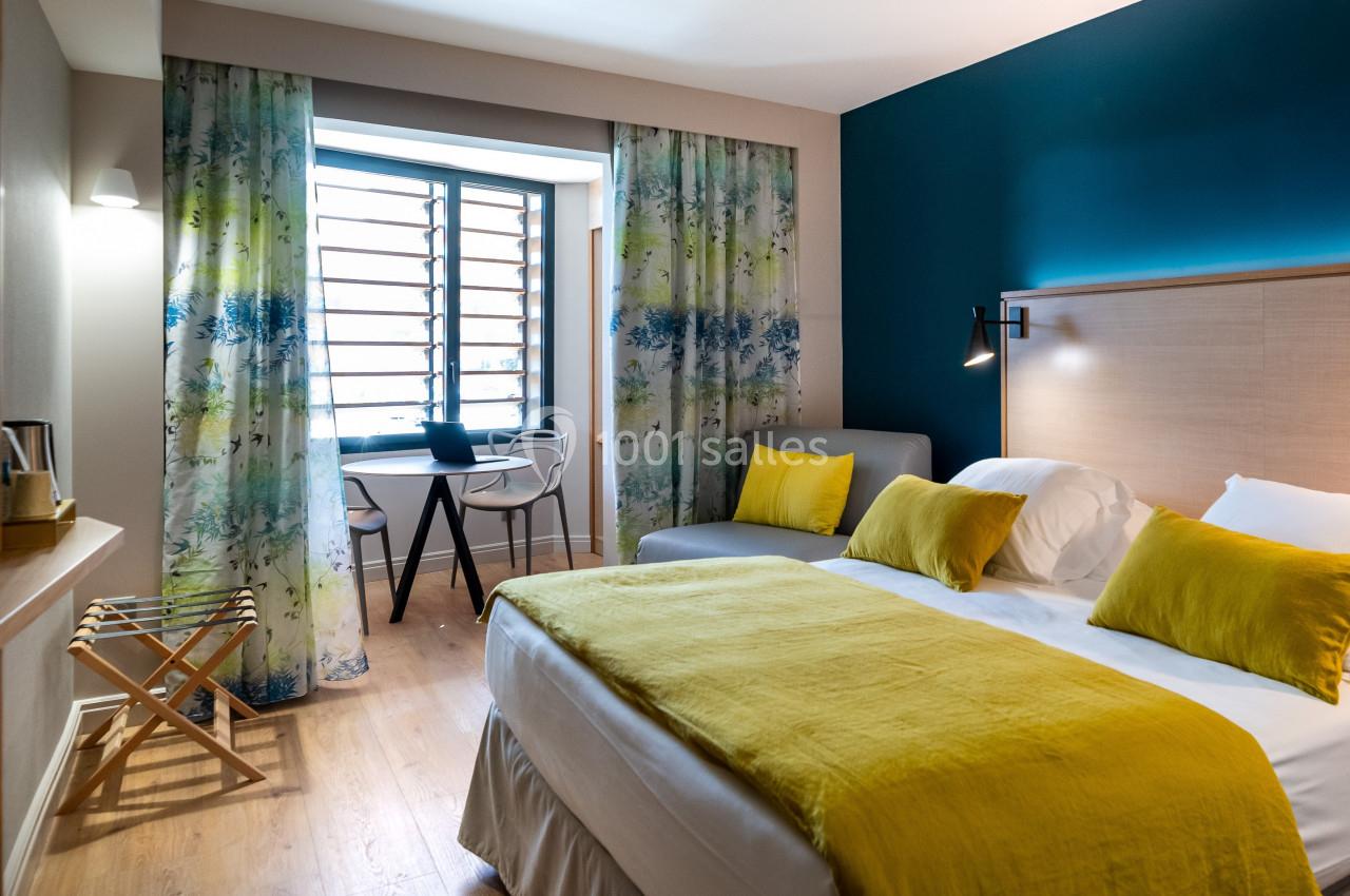 Chambre lumineuse avec lit double, coussins jaunes, bureau près de la fenêtre et rideaux à motifs floraux.