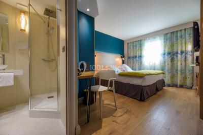 Salle lumineuse avec table en bois, chaises modernes, coin salon, lit double et murs aux tons neutres et bleu.