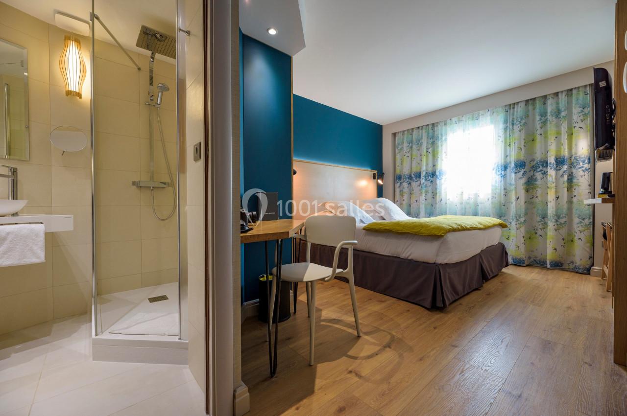 Chambre d'hôtel lumineuse avec lit double, bureau en bois, salle de bain attenante et grande fenêtre avec rideaux colorés.