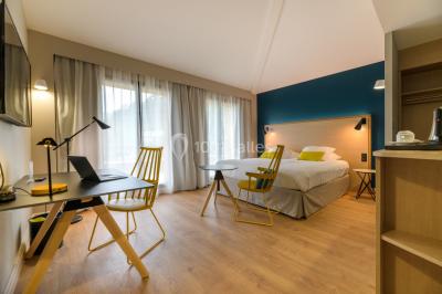 Salle lumineuse avec table en bois, chaises modernes, coin salon, lit double et murs aux tons neutres et bleu.