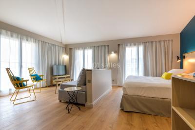 Salle lumineuse avec table en bois, chaises modernes, coin salon, lit double et murs aux tons neutres et bleu.