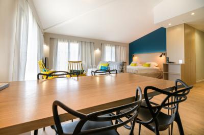 Salle lumineuse avec table en bois, chaises modernes, coin salon, lit double et murs aux tons neutres et bleu.