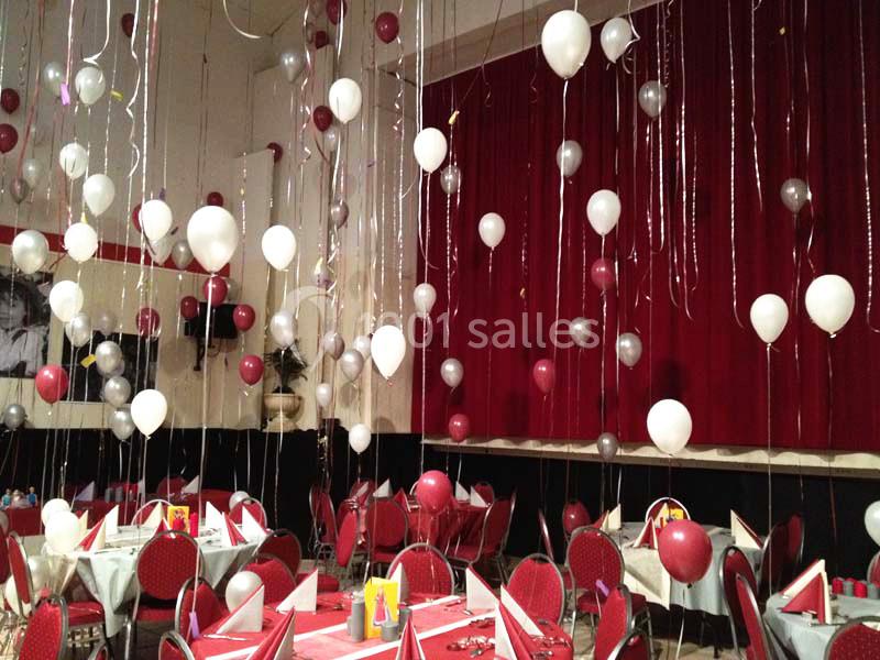 Salle décorée pour un événement avec des ballons rouges, blancs et argentés suspendus au plafond et tables dressées.